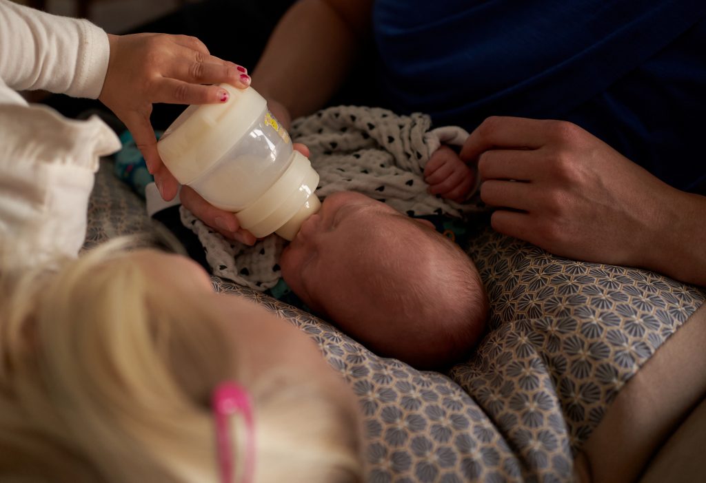 Geschwisterkind füttert Säugling im Wochenbett mit der Flasche und liegt kuschelnd neben Baby, während ein Elternteil auf der anderen Seite neben dem Baby kuschelt und liebevoll über das Babyhändchen streicht. Das Bild zeigt einen Ausschnitt eines Neugeborenenshootings im Wochenbett mit Geschwisterkind in Liechtenstein.